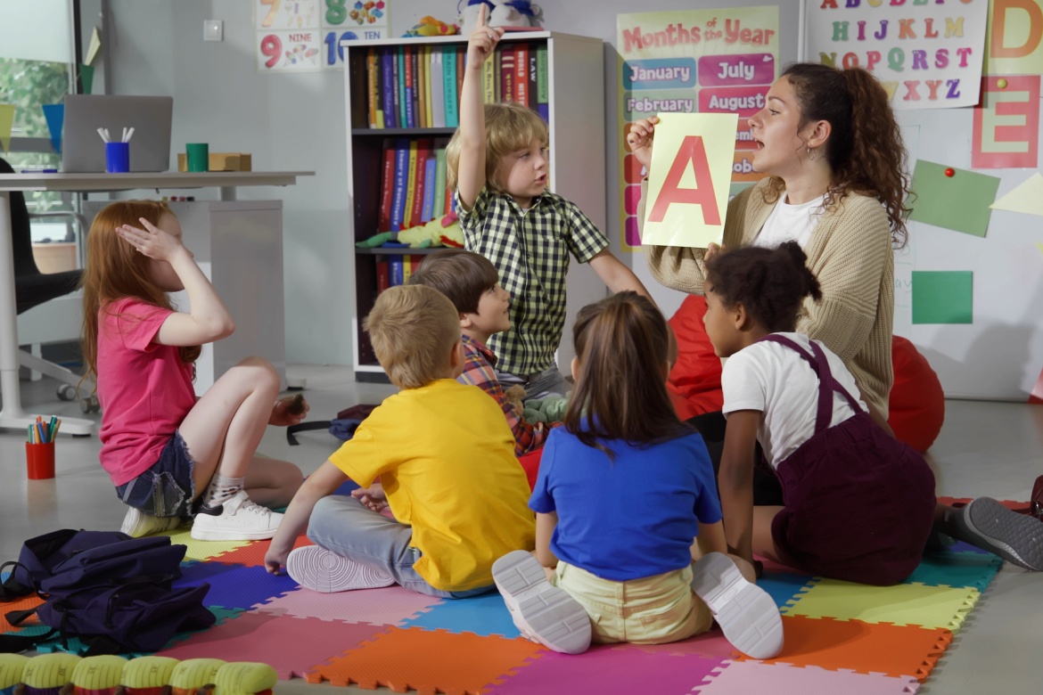 Female teacher hold flashcard with letter a teaching preschool kids alphabet. Diverse pupils of primary school sit on floor with teacher having alphabet lesson. Teacher without a degree 