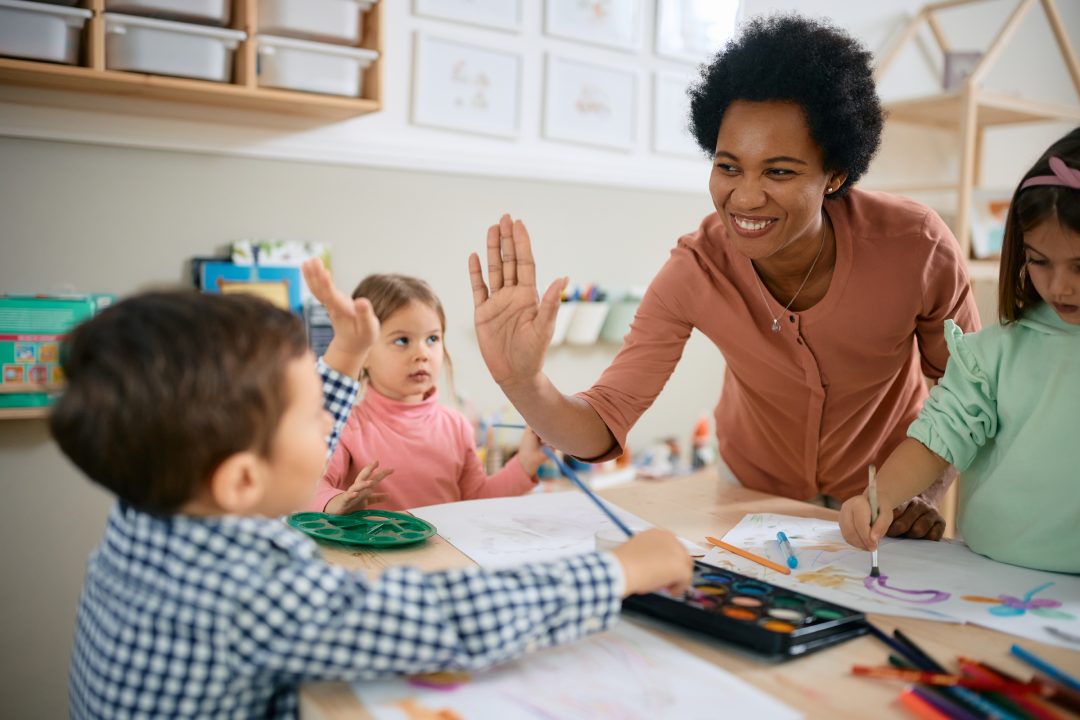 Maestra afroamericana feliz y niño pequeño chocando los cinco durante la clase de arte en el jardín de infancia.