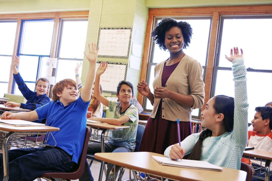 Escuela, tutor y alumnos levantan la mano para hacer o responder una pregunta académica para el aprendizaje. Diversidad, educación y niños de primaria hablando con su profesora en el aula.