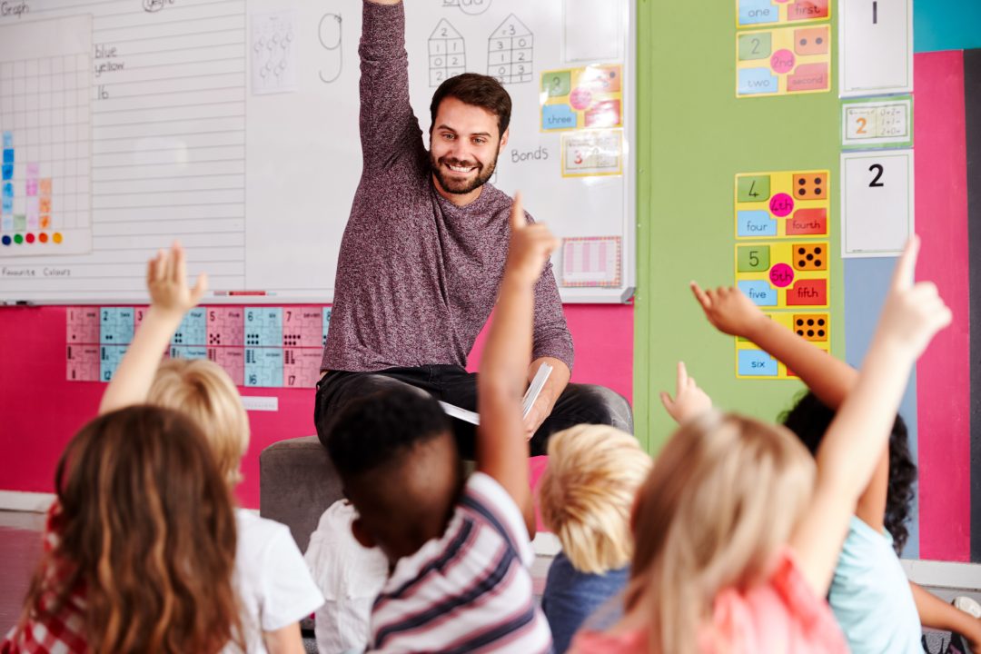 Elementary Pupils Raising Hands To Answer Question As Male Teacher Reads Story In Classroom