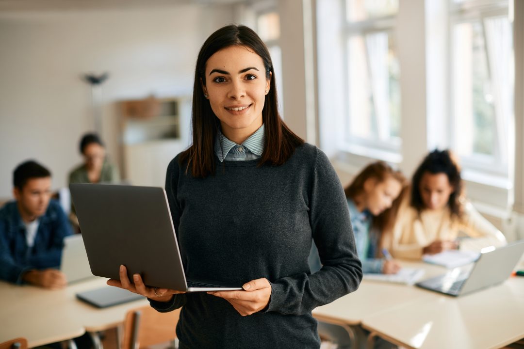 Profesora sonriente utilizando un ordenador portátil durante una clase de informática en un instituto y mirando a cámara. Sus alumnos están aprendiendo en el fondo.