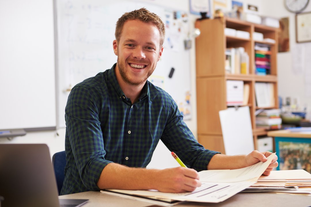 Happy bearded male teacher at desk looking to camera