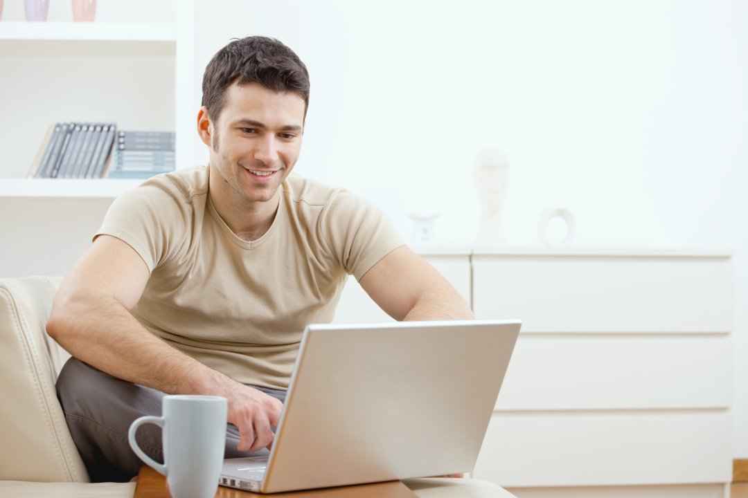 Happy young man in t-shirt sitting on sofa at home, working on laptop computer, smiling.
