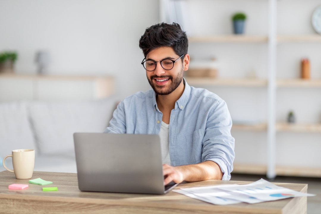 Retrato de hombre árabe feliz freelancer sentado en el escritorio con el ordenador portátil en la oficina en casa, mirando y sonriendo a la pantalla, el espacio de copia. Joven emprendedor disfrutando de trabajo a distancia