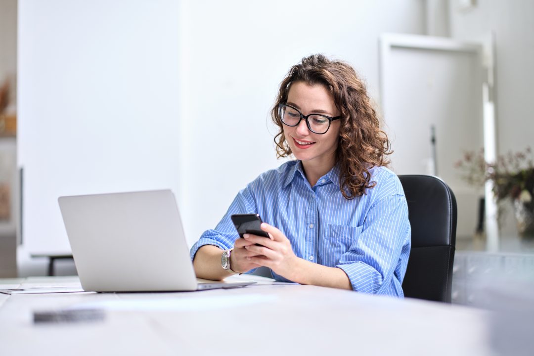 Young happy business woman, smiling pretty professional businesswoman worker looking at smartphone using cellphone mobile technology working at home or in office checking cell phone sitting at desk.