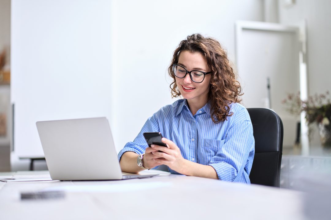 Joven mujer de negocios feliz, sonriendo bastante profesional mujer de negocios trabajadora mirando el teléfono inteligente utilizando la tecnología móvil del teléfono móvil de trabajo en casa o en la oficina de la comprobación de teléfono celular sentado en su escritorio.