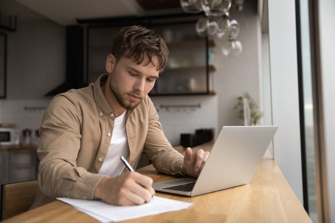 Joven estudiante preparar ensayo de investigación en la cocina moderna navegar por Internet utilizar el ordenador portátil escribir la información al informe de papel a mano. Focused man freelancer work from home make list of data from pc screen