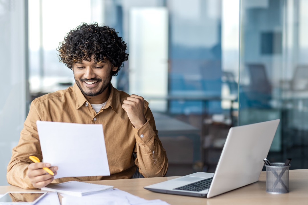 Witness the captivating essence of a vibrant Indian man with curly hair, fully immersed in his work as he sits at a table, surrounded by scattered papers, alongside his trusty laptop.