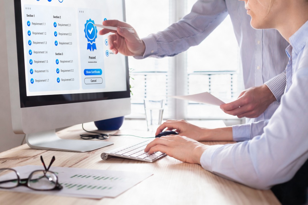 Gestión de la garantía y el control de calidad. Concepto de normalización y certificación. Cumplimiento de las normas ISO internacionales. Directivo y auditor mirando el certificado en la pantalla del ordenador.