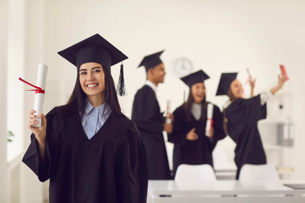 Graduada vestida con una toga de licenciada muestra a la cámara un diploma en sus manos mientras está de pie en el aula con el fondo de compañeros que se están haciendo un selfie. Concepto de educación.