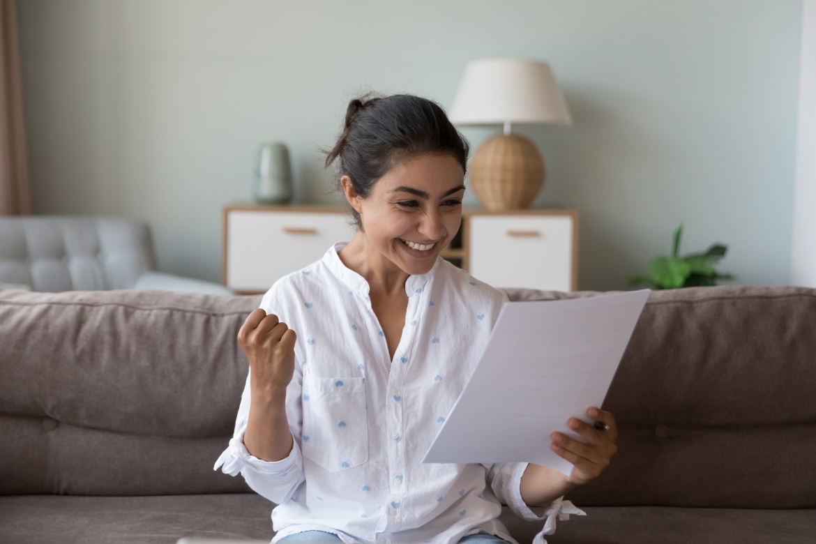 Joven india alegre y emocionada recibiendo una carta de buenas noticias, leyendo un documento en papel en casa, haciendo ganador sí mano, sintiéndose feliz, sonriendo, riendo, celebrando, éxito, lograr