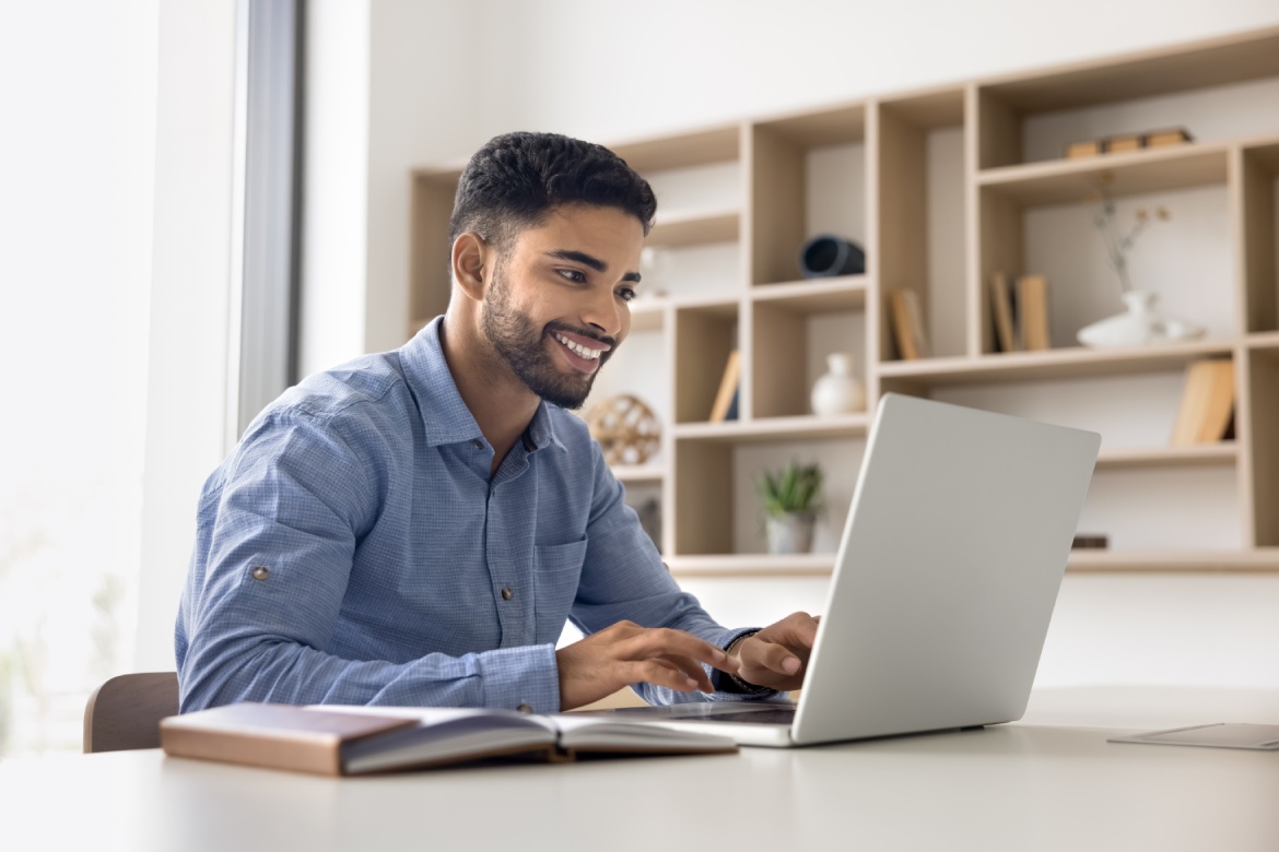 Hombre árabe joven satisfecho y positivo trabajando en la oficina solo, sentado en el lugar de trabajo con el ordenador escribiendo en el portátil, utilizando el servicio en línea en el PC para la comunicación de trabajo, sonriendo.