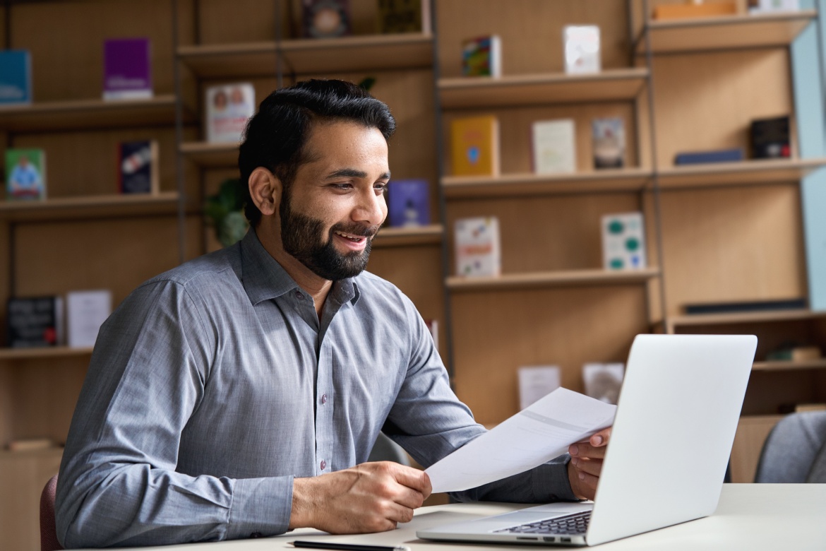 Hombre de negocios indio sonriente, empleador de recursos humanos, profesor o abogado con un documento de currículum vitae en una entrevista de trabajo virtual por videoconferencia, consultando a un cliente con un portátil en una asesoría jurídica en línea.