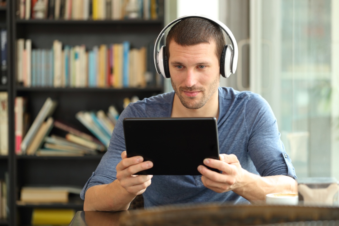 Hombre serio usando tableta y auriculares para ver medios online sentado en una cafetería
