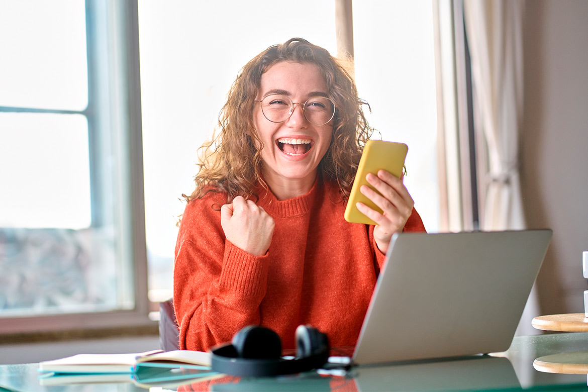 A woman at a table uses a laptop and phone, considering the possibility of teaching in Texas with a Nebraska certification.