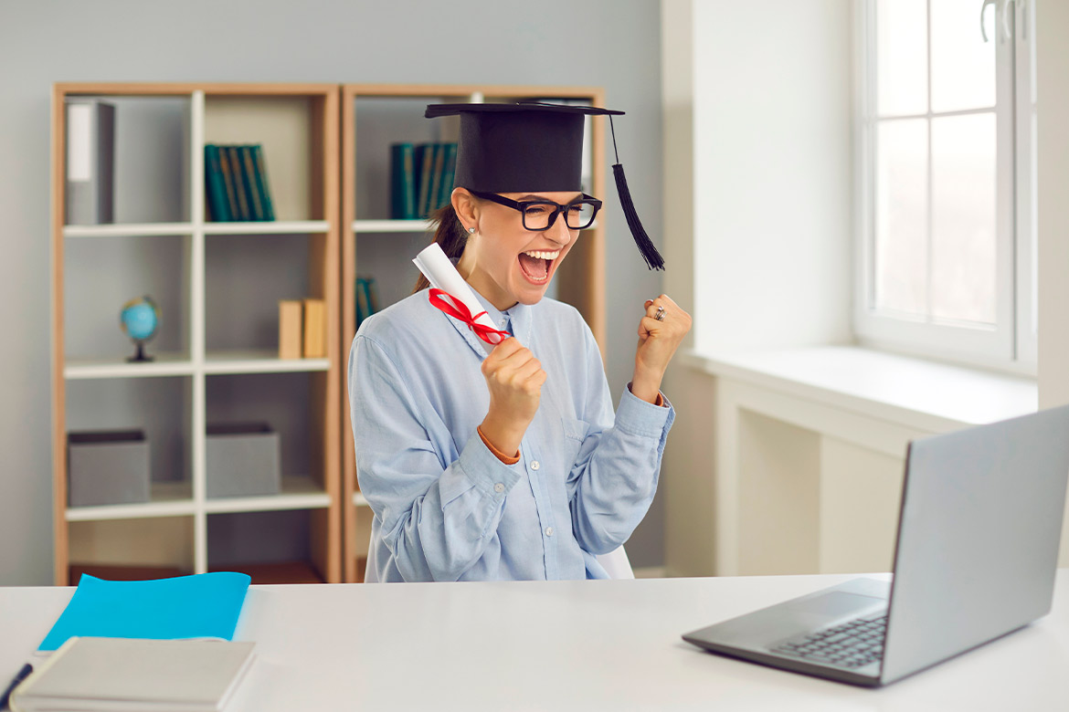A woman in a graduation cap and gown works at a desk with a laptop, highlighting her transition from student to educator.