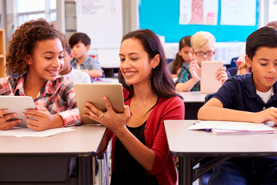 A classroom scene featuring a teacher and students interacting with a tablet, focused on educational tasks.
