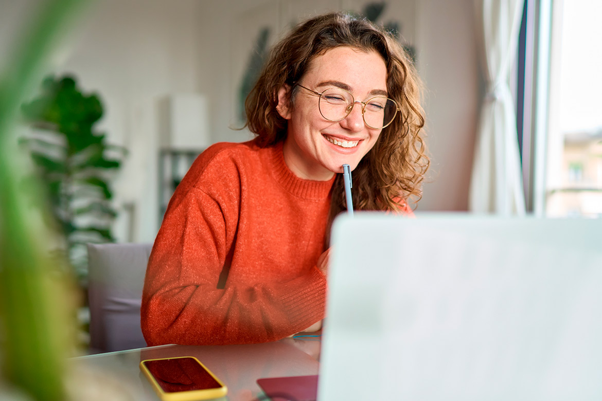 A smiling woman with glasses in a red sweater uses her laptop, exploring information on becoming a teacher in Lubbock.