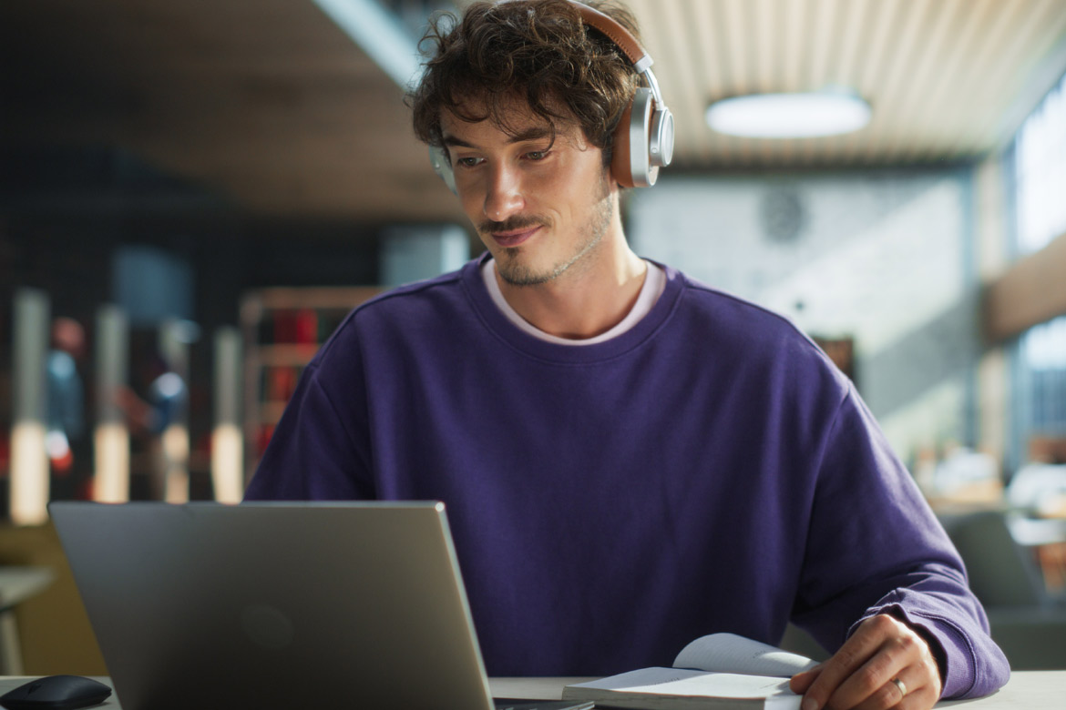 Handsome Student Writing an Essay on Laptop Computer, Thinking About the Topic, Brainstorming, Finding Solutions. Young Stylish Man Studying in a Public Library, Listening to Lecture in Headphones