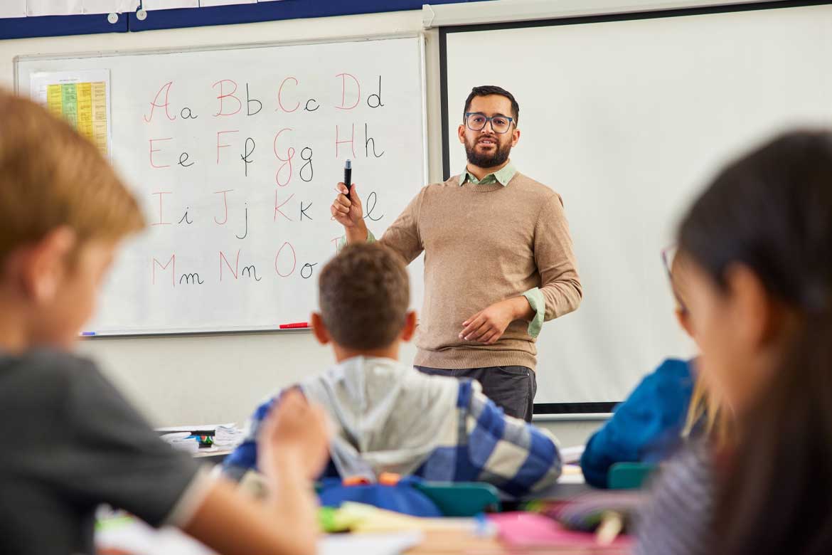 Smiling young male teacher helping students learn english alphabets in classroom. Rear view of elementary children revising and learning english letters at school. Male teacher teaching lesson.