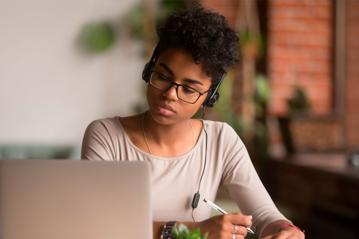 Focused multiracial woman wearing headphones watching webinar write notes study online with online teacher african female student learning language computer course on laptop listen translate lecture