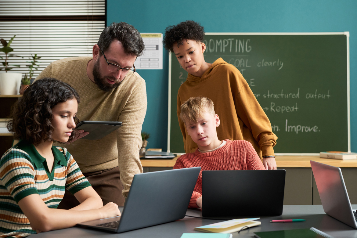 Caucasian man assisting diverse group of teenagers working on laptops in classroom, students focusing on screens while teacher holding digital tablet and explaining task