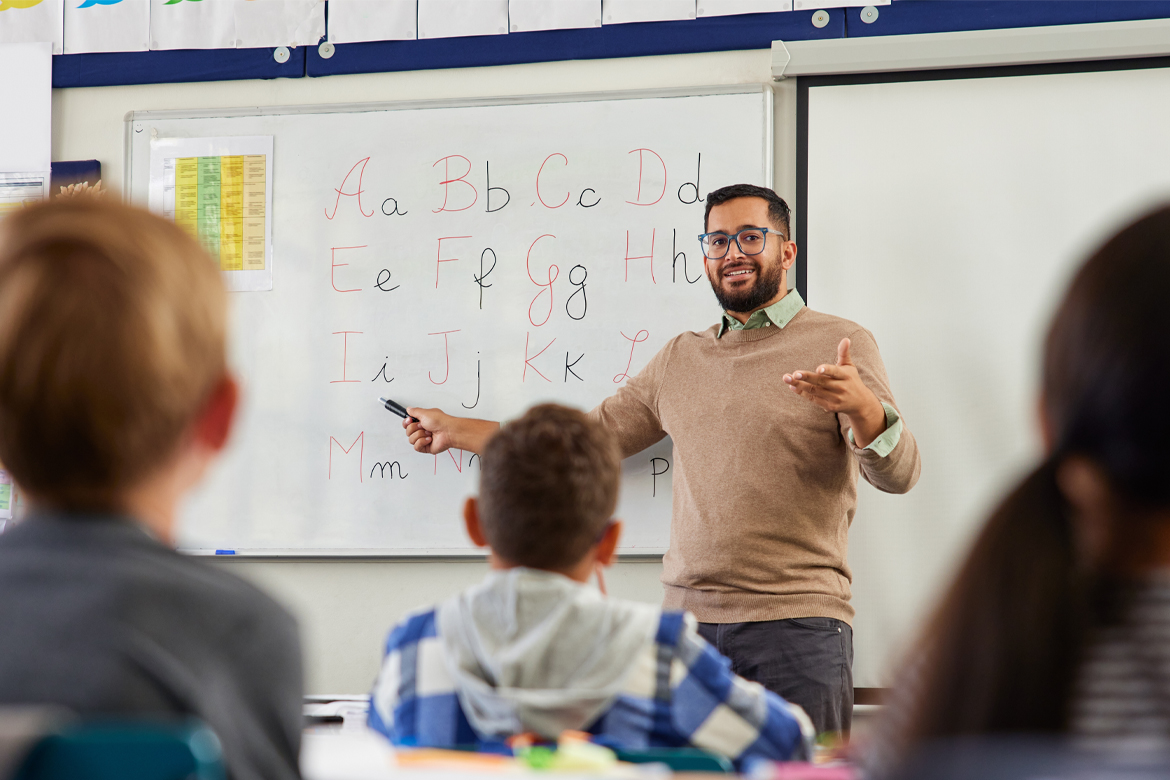 Smiling young male teacher helping students learn alphabet in first grader classroom. Rear view of children revising and learning letters in school classroom. Male teacher teaching alphabet to kids.